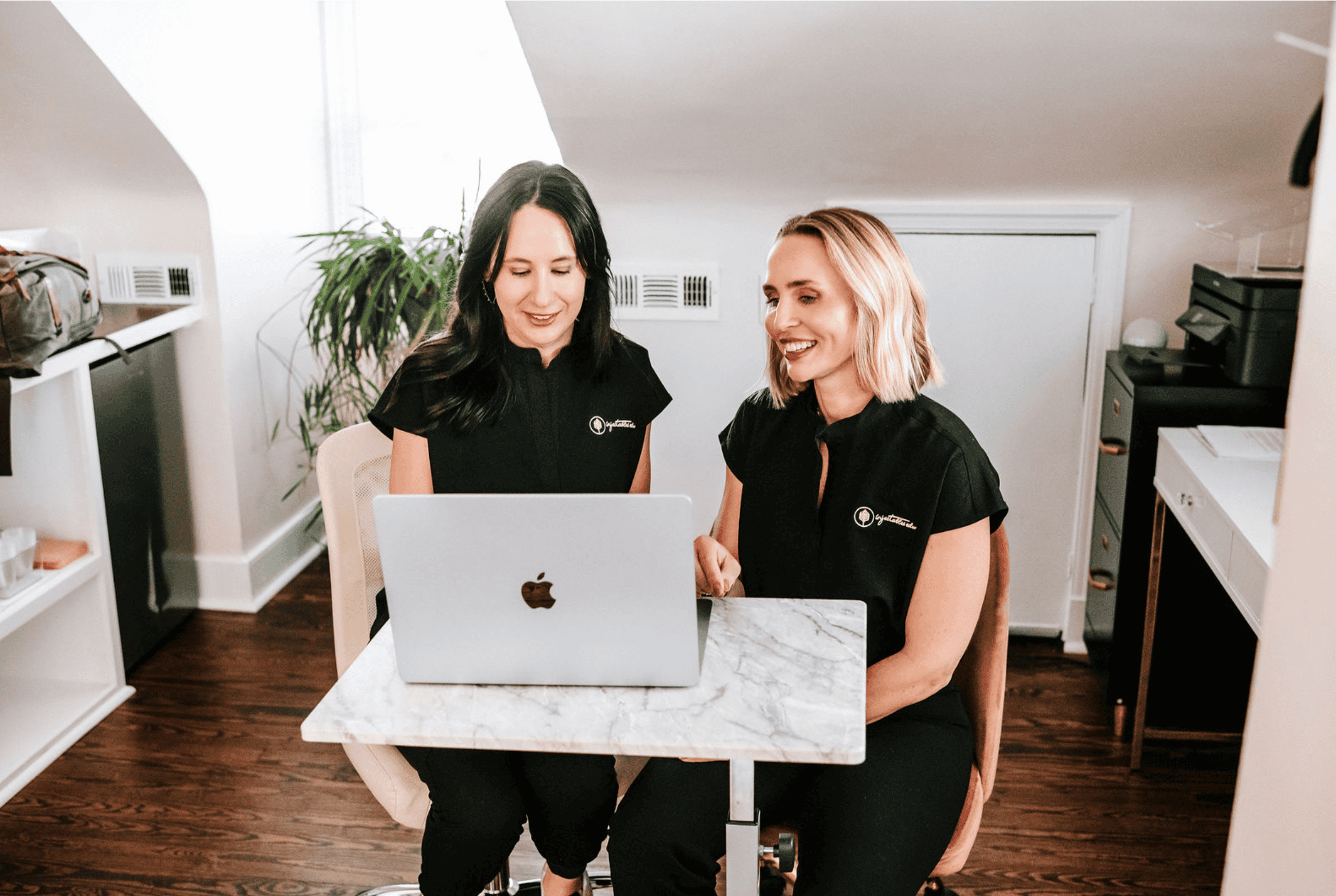 Two instructors from Injectables EDU smiling and reviewing training materials on a laptop.